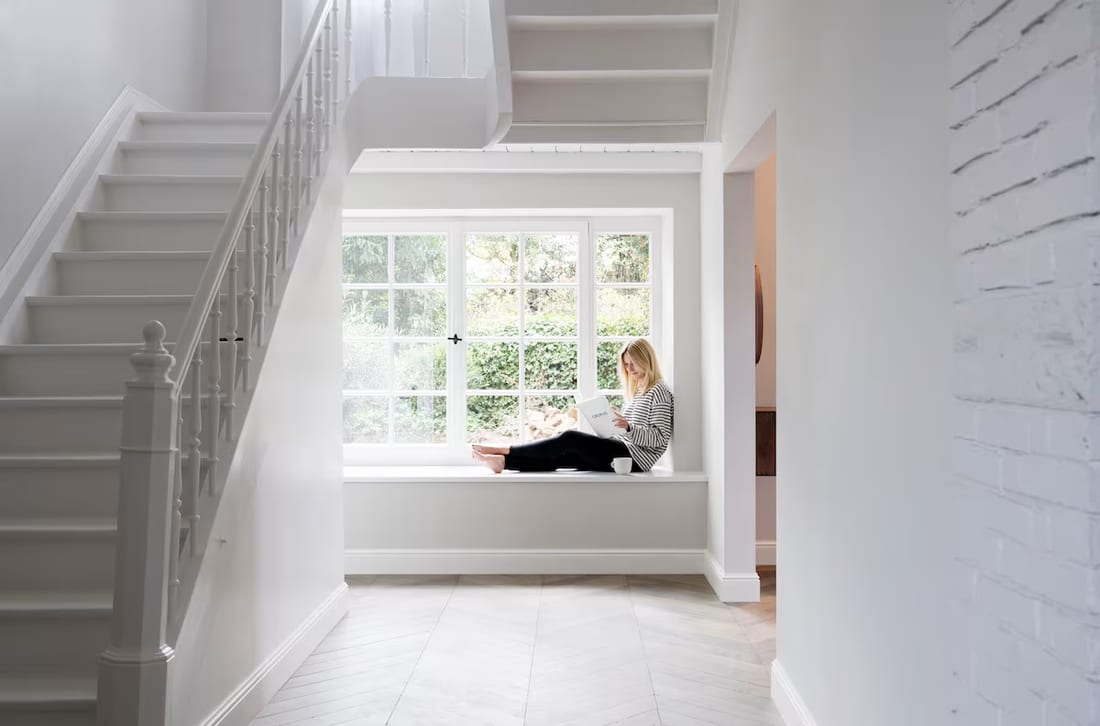 A woman reading in a sunlit window seat beside a staircase